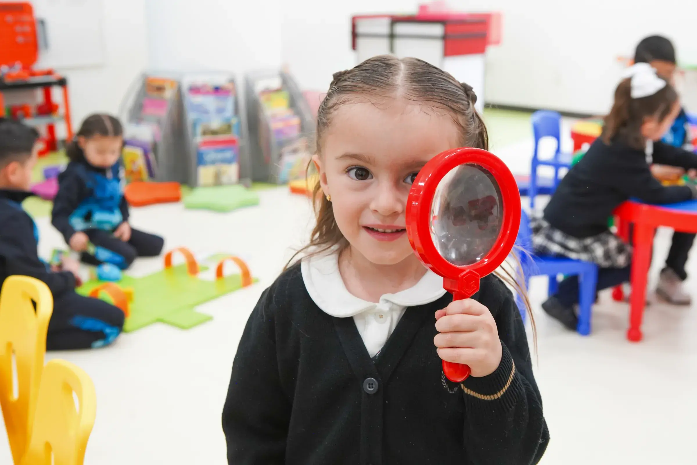 Niña en aula usando lupa roja durante actividad de exploración y descubrimiento en clase